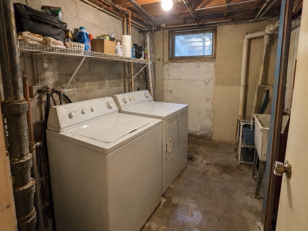 A cramped, unfinished laundry area with exposed pipes, bare concrete, and wire shelving